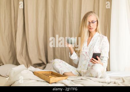 Young woman in eyeglasses checking news in her smartphone in bed during breakfast. Freelancer morning. Stock Photo