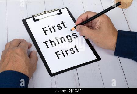 Hand with pencil writing the words First Things First on a paper with black clipboard to remind you an important task or appointment. Stock Photo
