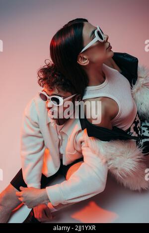 high angle view of stylish man in sunglasses and woman in faux fur jacket sitting on pink and grey Stock Photo