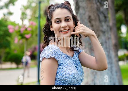 Young brunette girl smiling happy wearing summer dress on city park, outdoors makes a phone gesture and laughs looking at the camera. Stock Photo