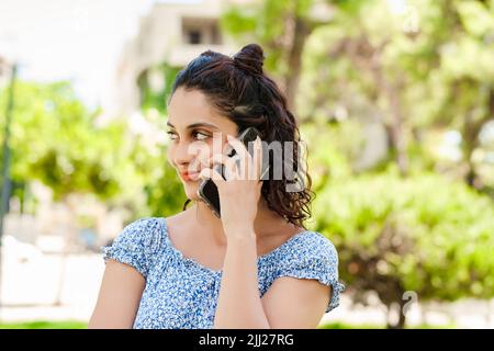 Beautiful brunette woman wearing summer dress standing on city park, outdoors talking on mobile phone with friends or boyfriend with smiles. Stock Photo