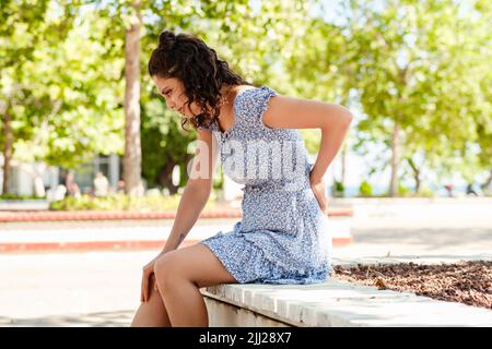 Young brunette woman wearing summer dress on city park, outdoors back pain suffer from lower lumbar discomfort muscle pain. Massaging tense muscles. Stock Photo