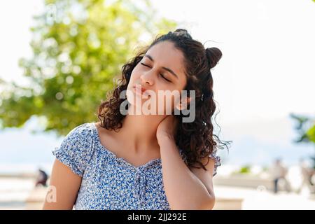 Portrait of young brunette woman wearing summer dress standing on city park, outdoors feeling hurt joint shoulder back pain ache, fibromyalgia concept Stock Photo