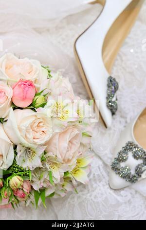 Wedding composition with silk white shoes, pastel bridal bouquet on white lace blurry background. Top view Stock Photo