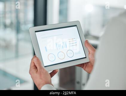 How are we doing this month, business wise. an unrecognisable businessman standing alone in his office and using a digital tablet. Stock Photo