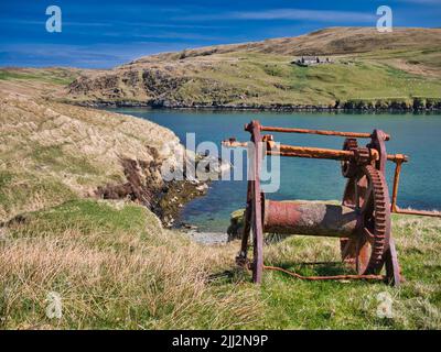 An abandoned, rusting boat winch with beach below in Shetland, UK, taken on a sunny day with blue sea in the background. Stock Photo