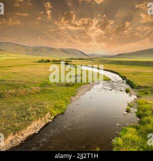 The small river flows towards the hills at sunset. Stream flowing through meadows. Stock Photo