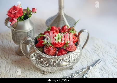 Strawberries in a silver vase on the table in a romantic style. Still life of strawberries, a bouquet of roses and a jug. Stock Photo