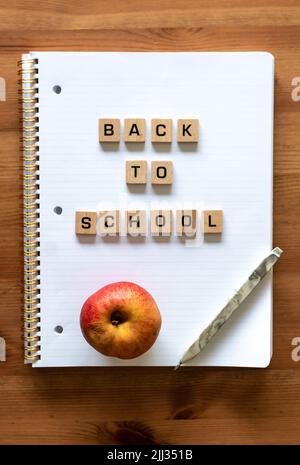 Wooden letters spell out 'back to school' on a blank white lined notepad beside a red apple and a pen. Hardwood desk surface. Learning. Stock Photo