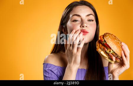 Chewing woman holding tasty fast food burger, cleaning her mouth after taking a bite. Satisfied girl eats her delivery meal, orange background. Stock Photo