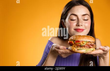 Smiling woman smells her tasty fresh burger from restaurant delivery. Girl holds cheeseburger on plate and sniff it, stands over orange background Stock Photo