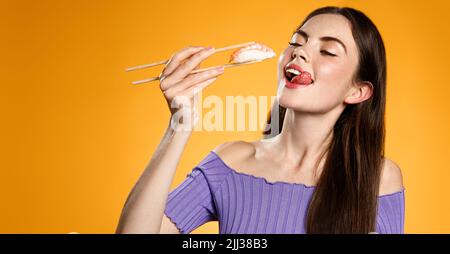 Hungry beautiful girl eats sushi with chopsticks, enjoys fresh nigiri in asian restaurant, orange background Stock Photo