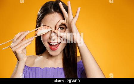 Asian restaurant and food delivery concept. Happy woman holds sushi nigiri with chopsticks, shows okay eye sign, orange background Stock Photo