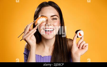 Happy woman with asian food. Girl with chopsticks eats sushi rolls and nigiri, order take out delivery, orange background Stock Photo