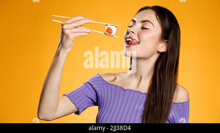Hungry girl eats sushi roll from chopsticks, takes bite of makizushi from asian restaurant delivery, orange background Stock Photo
