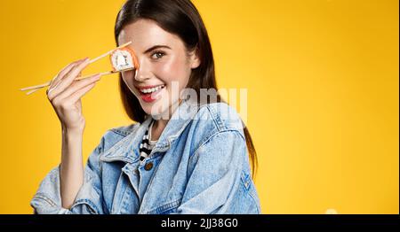 Beautiful modern girl posing with sushi roll and chopsticks on orange background Stock Photo