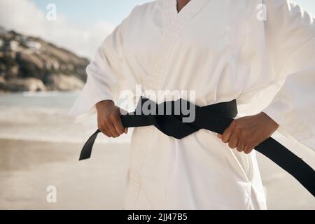 Shes right at the top of the game. an unrecognizable female martial artist tying her black belt while practicing karate on the beach. Stock Photo