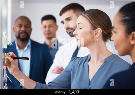 Confident manager showing and teaching team of colleagues while writing on sticky notes on transparent board in office. Diverse group of business Stock Photo