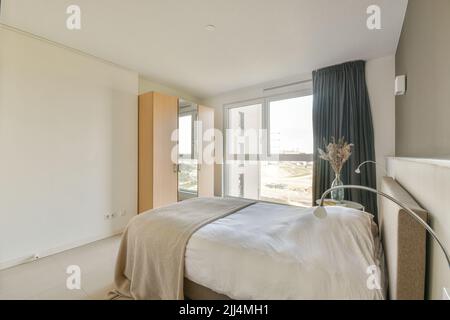 Retro bedside table with books and lamp located near comfortable bed and shelf with decorations against window in modern bedroom Stock Photo