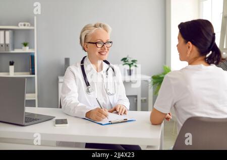 Happy smiling doctor at clinic talking to woman, giving consultation and taking notes Stock Photo