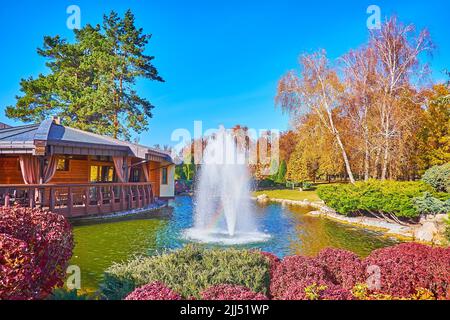 The picturesque autumn plants - red barberry bushes, green juniper, yellow birches and a fountain on the lake with bright rainbow in front of it, Mezh Stock Photo