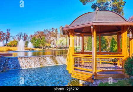 The beautiful wooden gazebo on the lake with small waterfall and fountains in background, Mezhyhirya, Ukraine Stock Photo
