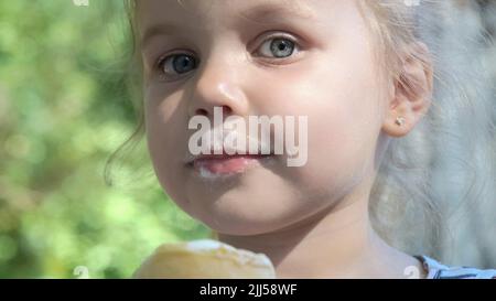 Cute little girl eats ice cream outside. Close-up portrait of blonde girl sitting on park bench and eating icecream. Stock Photo