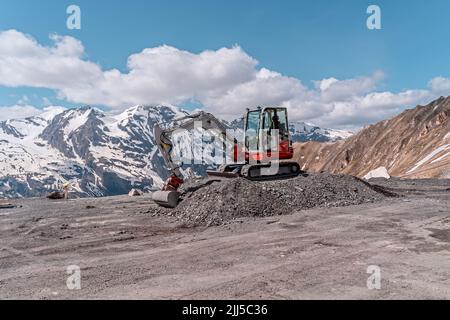 red excavator with shovel standing on hill with stones, mountains in background Stock Photo