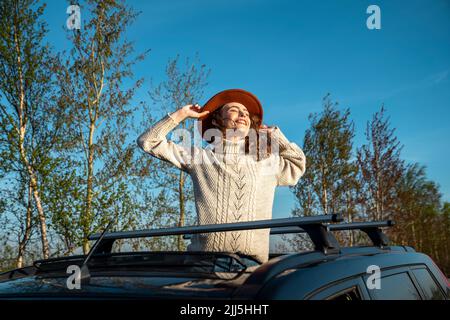 Happy woman wearing hat standing with eyes closed through sun roof of car Stock Photo