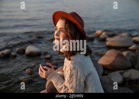 Smiling woman wearing hat sitting with coffee cup enjoying sunset Stock Photo