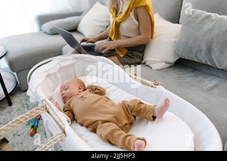 Woman using laptop sitting by baby sleeping in crib at home Stock Photo