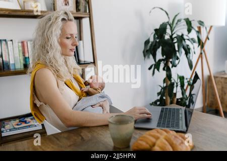 Mother carrying baby in arm using laptop at home Stock Photo