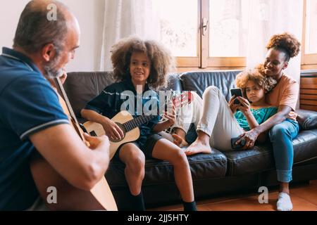 Man teaching son to play guitar sitting by woman and daughter on sofa at home Stock Photo