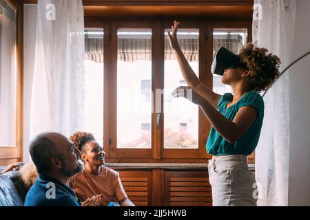 Girl wearing virtual reality simulator gesturing in front of family at home Stock Photo