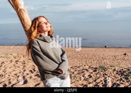 Young woman enjoying sunlight leaning on tree at beach Stock Photo