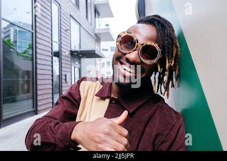 Happy man wearing sunglasses making thumbs up gesture leaning on wall Stock Photo