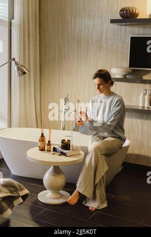 Woman using smart phone sitting on bathtub in bathroom Stock Photo