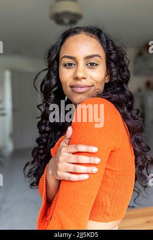 Happy woman with curly hair touching arm at home Stock Photo