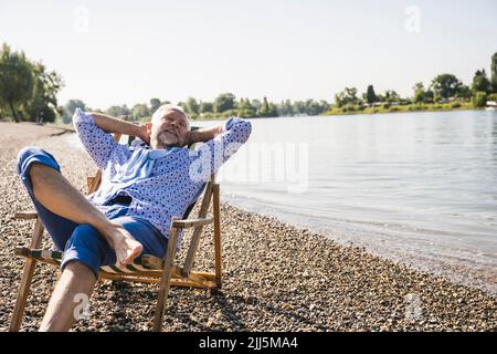 Smiling man with hands behind head resting on deck chair at riverbank Stock Photo