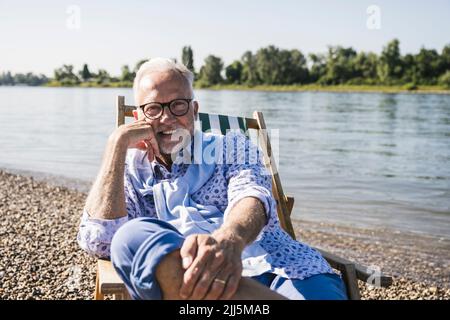 Smiling man wearing eyeglasses sitting on deck chair at riverbank Stock Photo