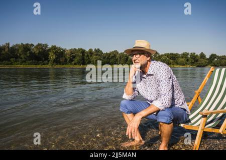 Man wearing hat sitting on deck chair at riverbank Stock Photo