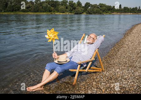 Senior man holding paper pinwheel toy sitting on deck chair at riverbank Stock Photo