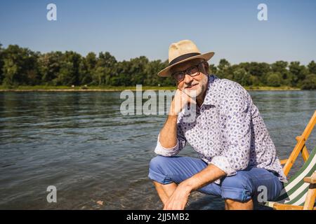 Smiling man wearing hat sitting on deck chair at riverbank Stock Photo