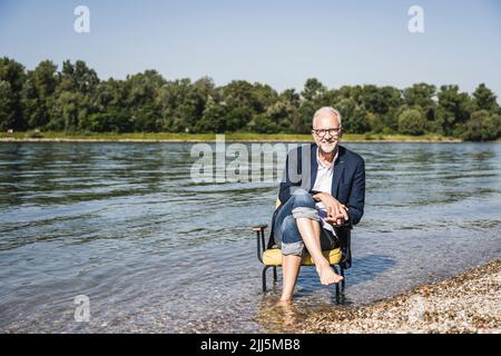 Smiling man wearing blazer sitting on chair at riverbank Stock Photo