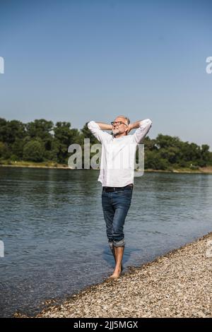 Senior man standing with hands behind head at riverbank Stock Photo