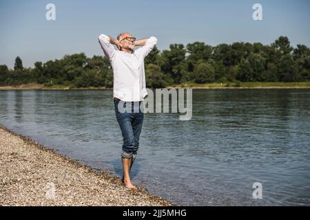 Smiling man standing with hands behind head at riverbank Stock Photo