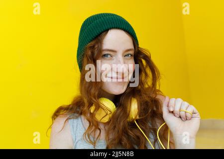 Smiling woman with headphones wearing green knit hat in front of yellow wall Stock Photo