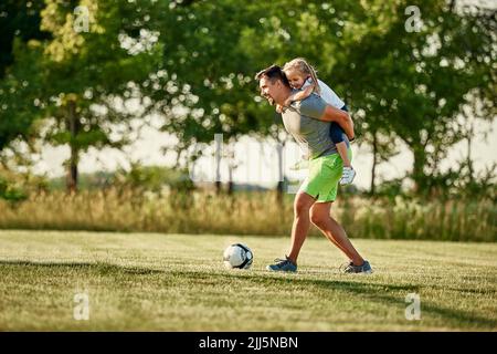 Happy girl enjoying piggyback ride given by man playing soccer at sports field Stock Photo