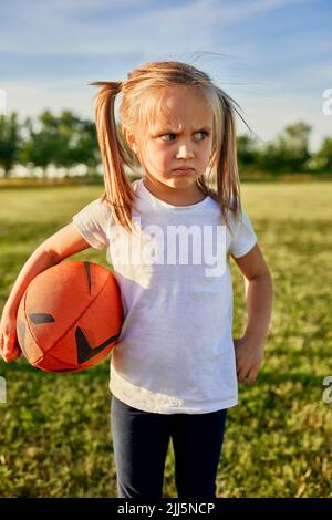 Displeased blond girl with rugby ball standing at sports field on sunny day Stock Photo