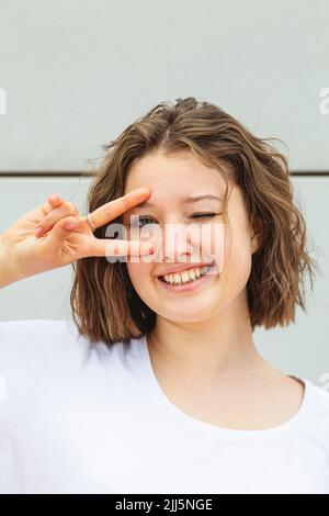 Happy teenage girl winking eye gesturing peace sign in front of wall Stock Photo